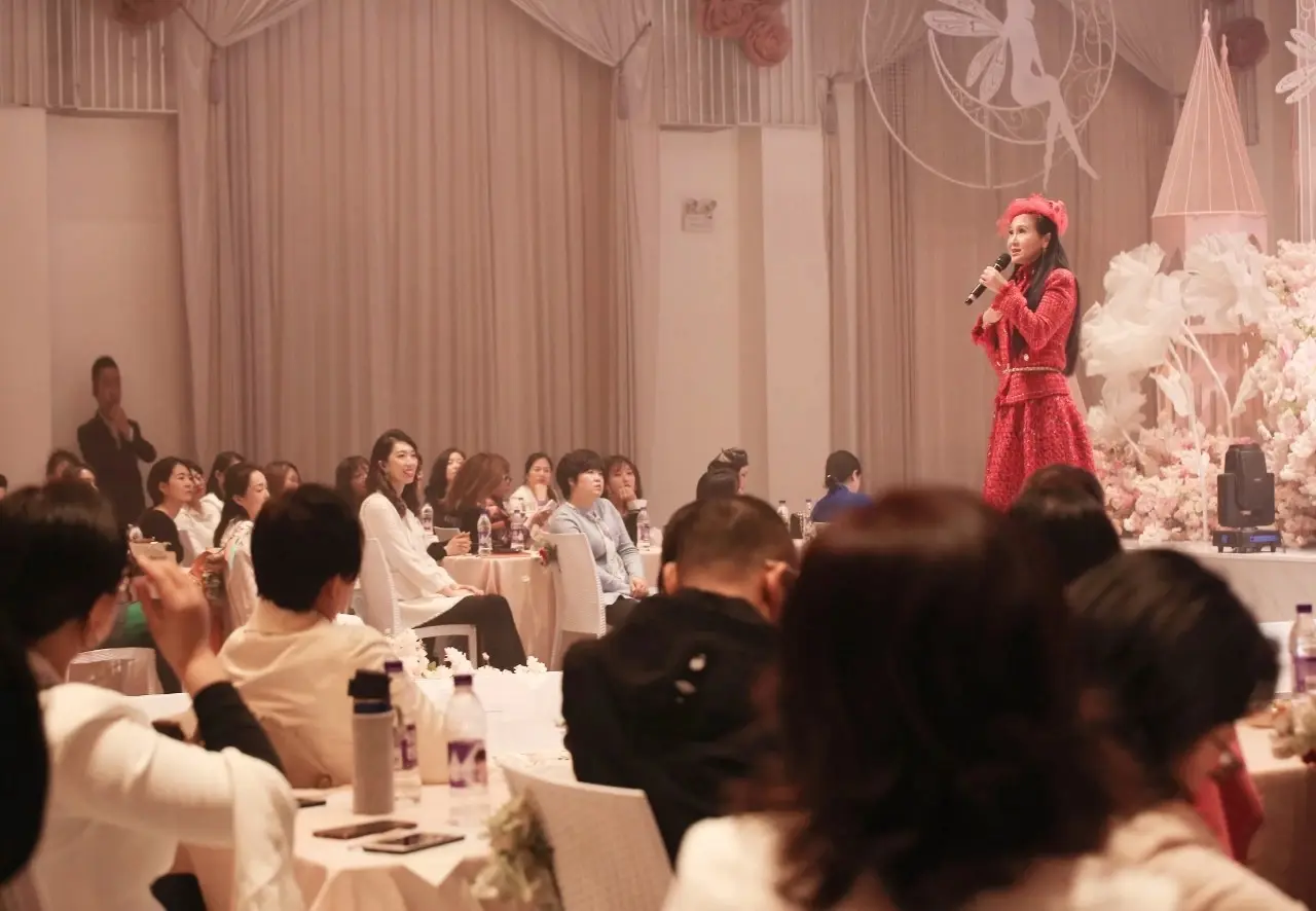 Jessica Chaijaya, en tenue rouge et chapeau rouge, parle au micro devant un public assis dans une salle de banquet