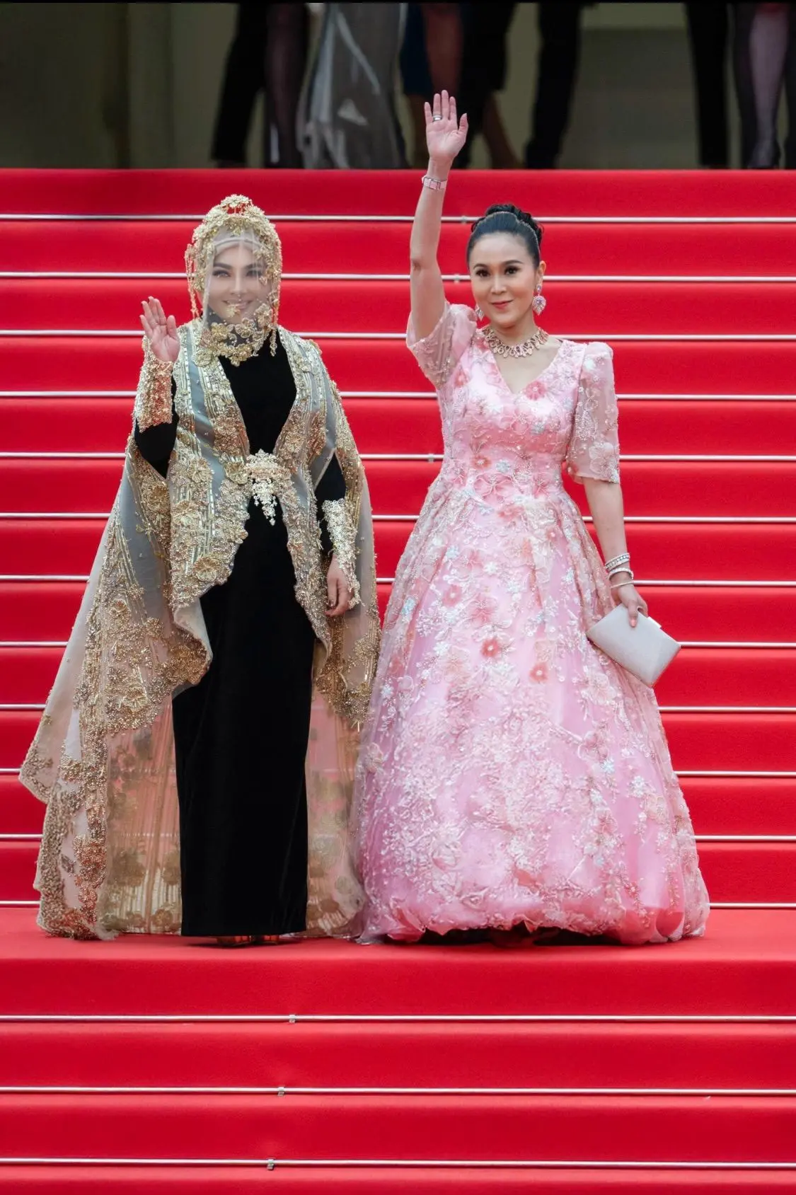 Syahrini, en robe noir et or avec une coiffe dorée, et Jessica Chaijaya, en robe rose, saluent depuis des marches recouvertes du tapis rouge