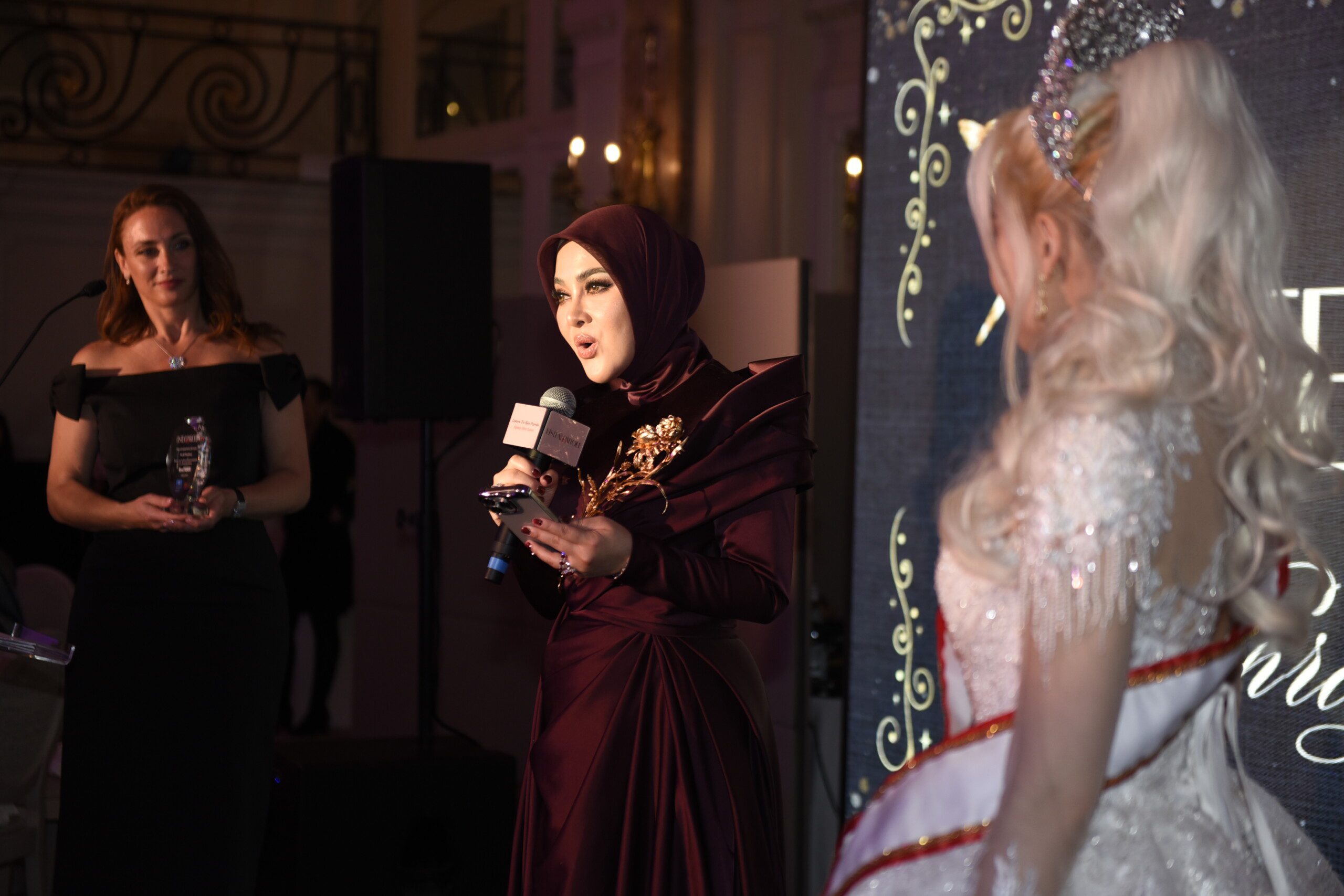 印尼巨星茜拉莉妮在2025戛纳电影节慈善晚宴演讲台照片：她身着晚礼服手持奖杯，身后是卡尔顿酒店大沙龙宴会厅的水晶吊灯与宾客。表情真挚，展现艺人与慈善家的双重身份。